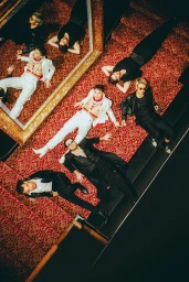 Group of men posed dramatically on a patterned red carpet staircase reflected in a large ornate mirror.