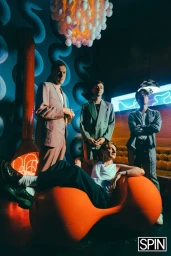 Four men posing in a brightly lit interior space with an orange modern sculptural chair, blue patterned walls, and a hanging globe installation.