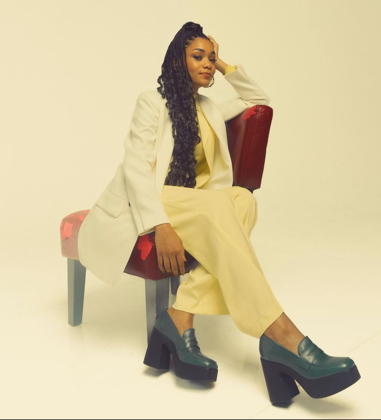 Woman in yellow pantsuit and white blazer poses on a modern stool against a white background, wearing block heel loafers.