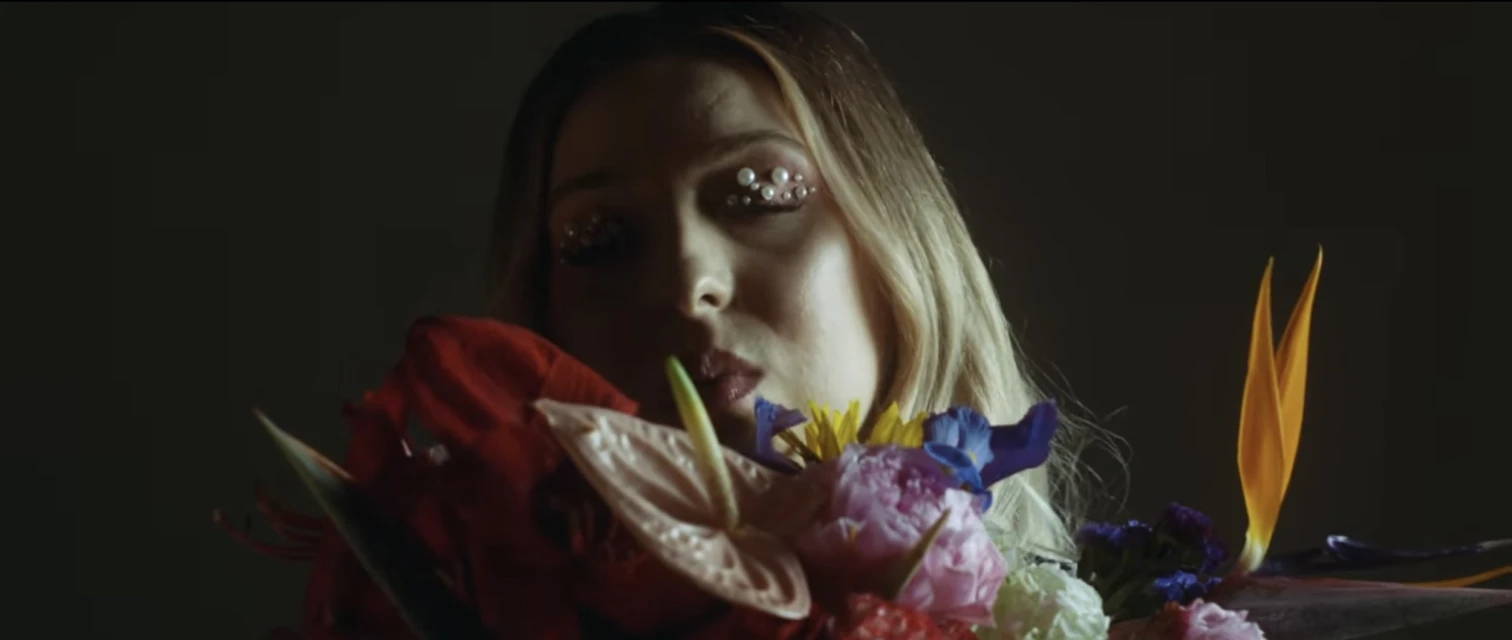 Portrait of a young woman with blond hair and pearls decorating her eyes, holding a vibrant bouquet of flowers in the foreground.