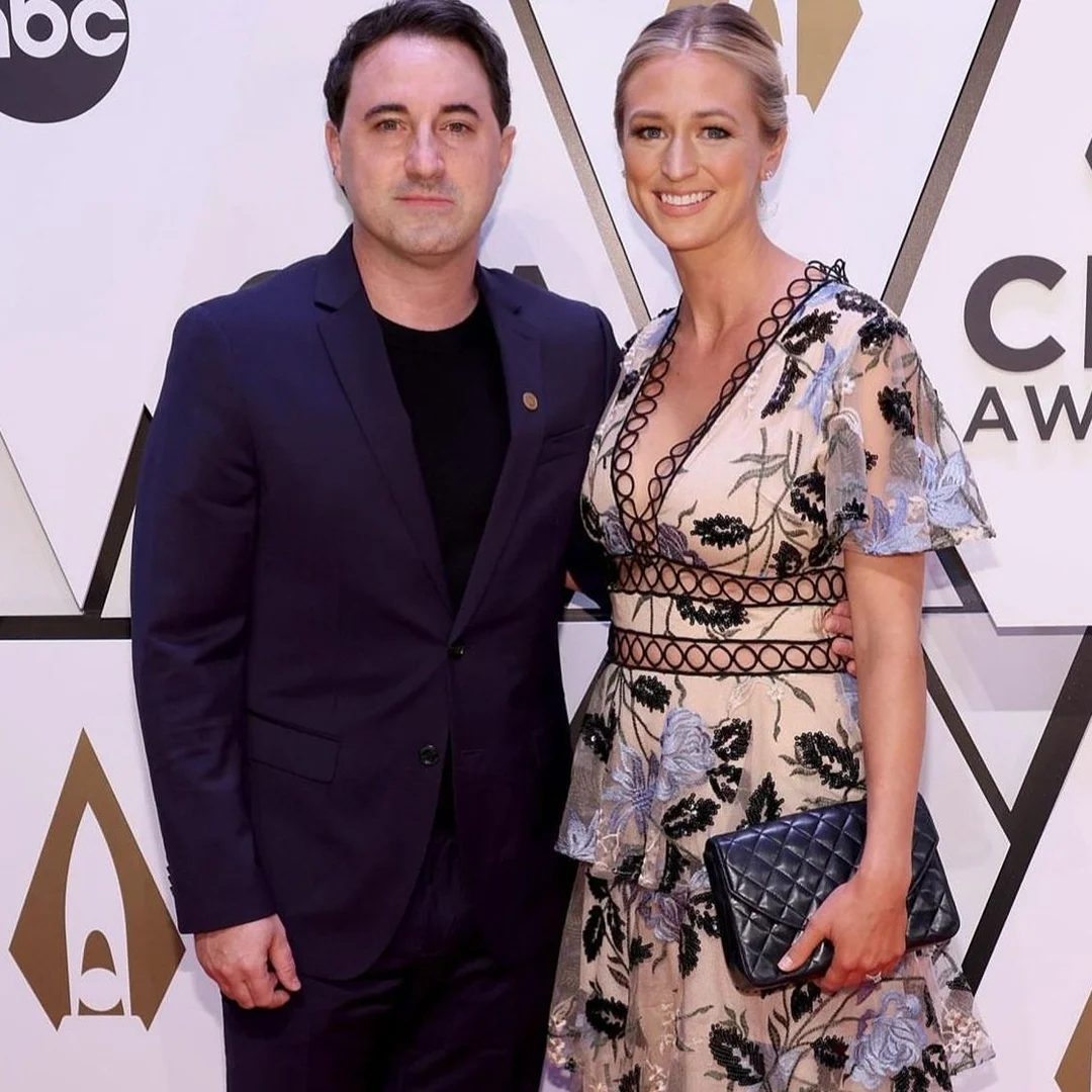 A smiling couple poses together on the red carpet. The woman is wearing a floral dress and carrying a clutch. The man is wearing a suit.