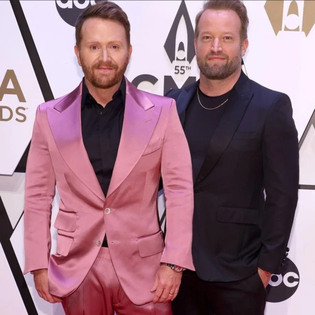 Two men stand closely on red carpet. One in pink suit, the other in dark suit. Event logo visible behind them.