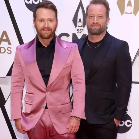 Two men stand closely on red carpet. One in pink suit, the other in dark suit. Event logo visible behind them.