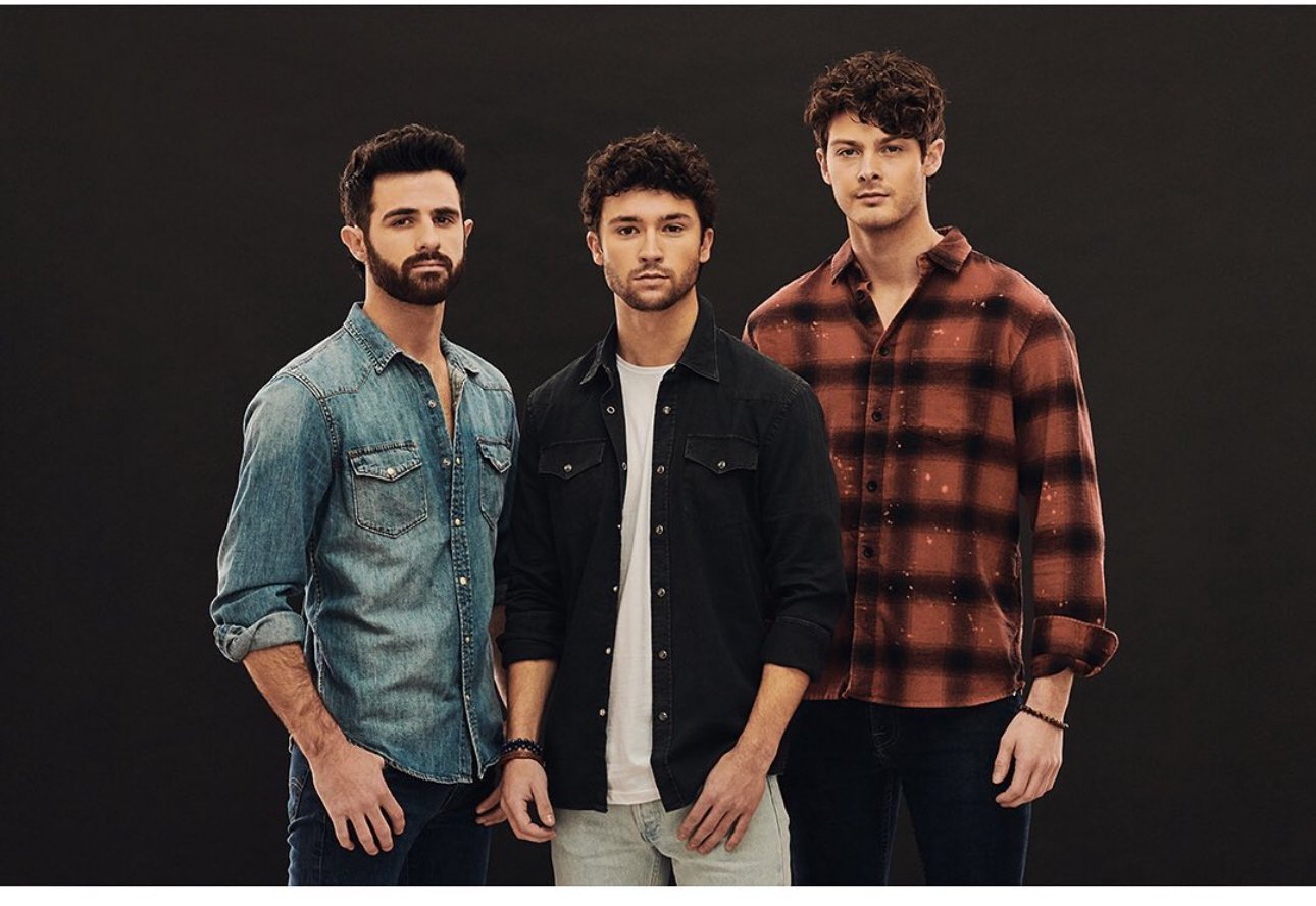 Three adult men stand in a studio wearing casual denim and flannel button-down shirts against a dark background in a medium shot.