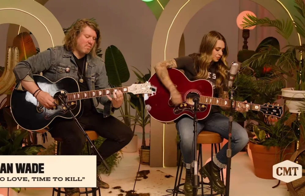 Two musicians, a man and a woman, play acoustic guitars in a warm-toned, plant-filled indoor setting with vintage microphone and CMT logo.