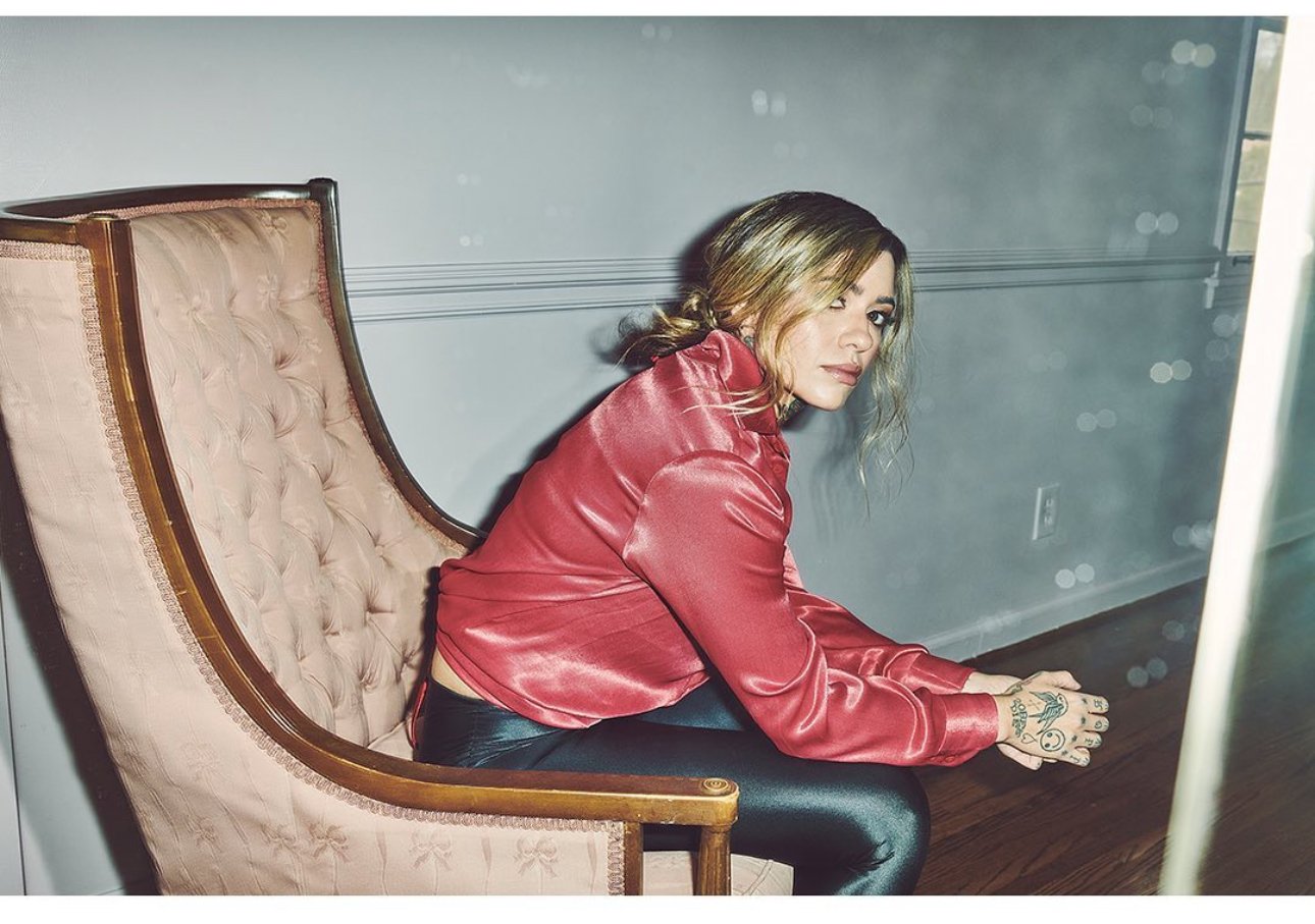Young woman in a rose blouse sits in a vintage chair indoors. She has tattoos, short hair, and is sitting in a relaxed pose.