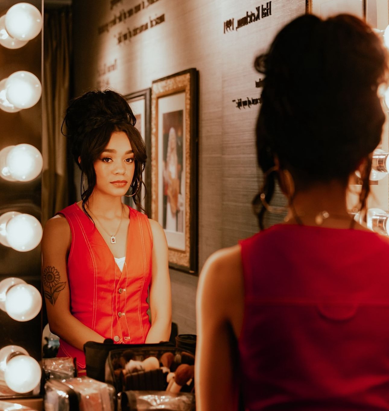 Young woman with sunflower tattoo poses in a red vest in front of a mirror with bulb lighting in an indoor dressing room.
