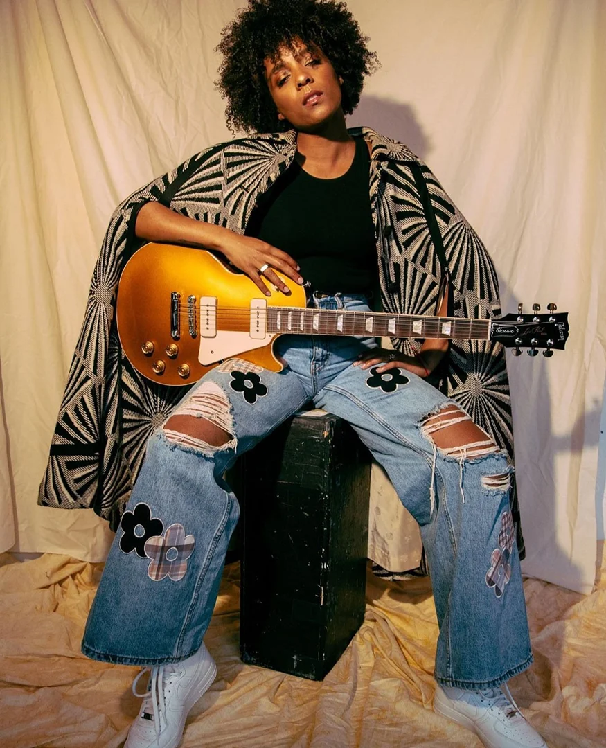 Black woman with voluminous curly hair holding an electric guitar while seated on a crate against a textured fabric backdrop.