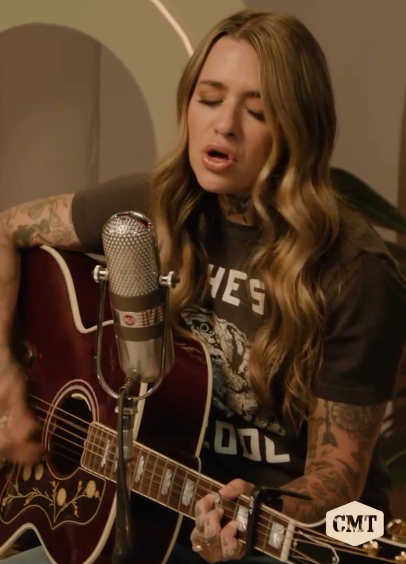 Blonde woman sings into a vintage microphone while playing an acoustic guitar with floral inlays and decorative details.
