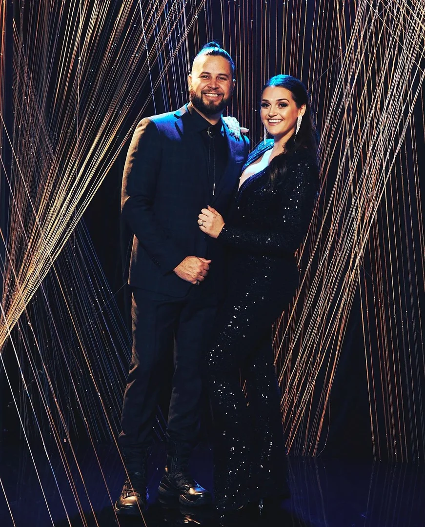 Elegant couple posing together in formal wear against a shimmering gold-toned backdrop with a dark atmosphere and soft, diffused lighting.