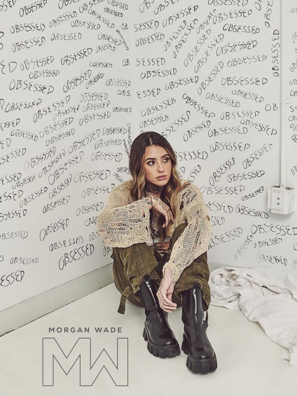 Morgan Wade poses in a beige knit sweater, cargo pants, and boots in a room with 'OBSESSED' graffiti. Close-up shot with Morgan Wade logo.