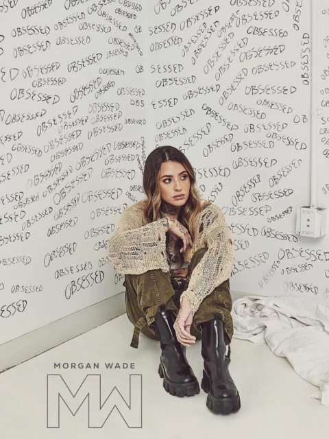 Morgan Wade poses in a beige knit sweater, cargo pants, and boots in a room with 'OBSESSED' graffiti. Close-up shot with Morgan Wade logo.