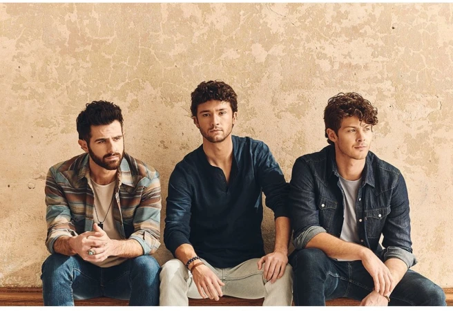 Three men seated against a weathered textured wall wearing casual shirts and denim jeans