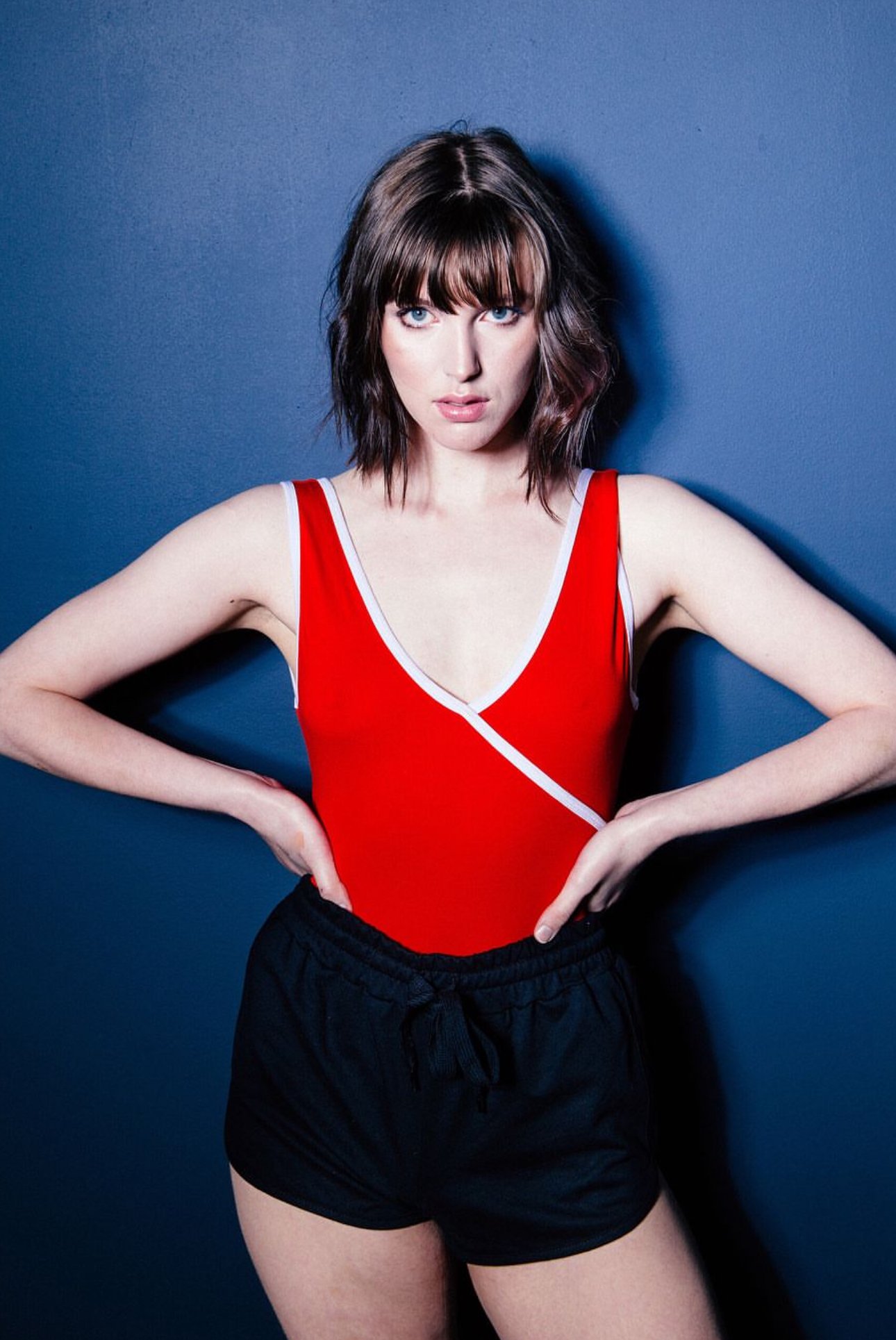 A young adult woman in a red one-piece swimsuit and black shorts poses against a dark blue background with hands on her hips.