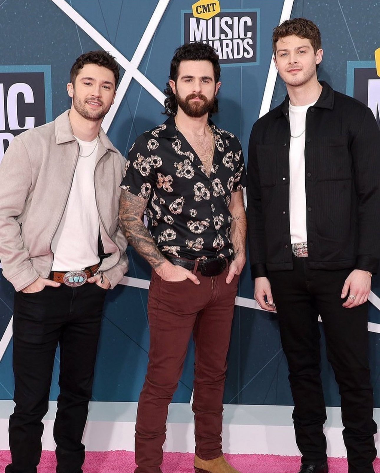 Three adult males pose on the CMT Music Awards pink carpet in front of a logo backdrop, dressed in casual fashionable outfits.