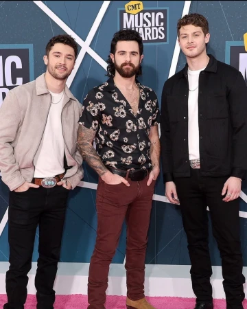 Three young adult men pose on a pink carpet at the CMT Music Awards, wearing trendy clothing and accessories.
