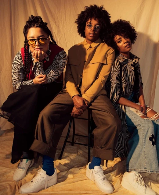 Three young adults pose sitting on a stool in a studio with warm lighting wearing retro-inspired outfits and fashionable sneakers.