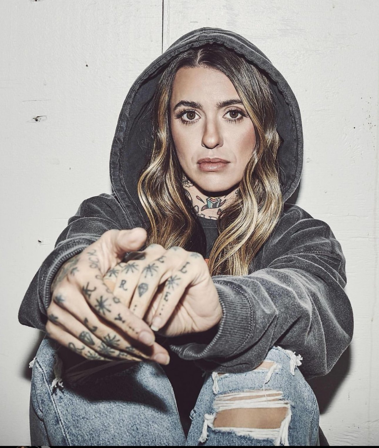 Portrait of a young adult woman with tattoos, wearing a hooded sweatshirt and ripped jeans, indoors against a neutral background.