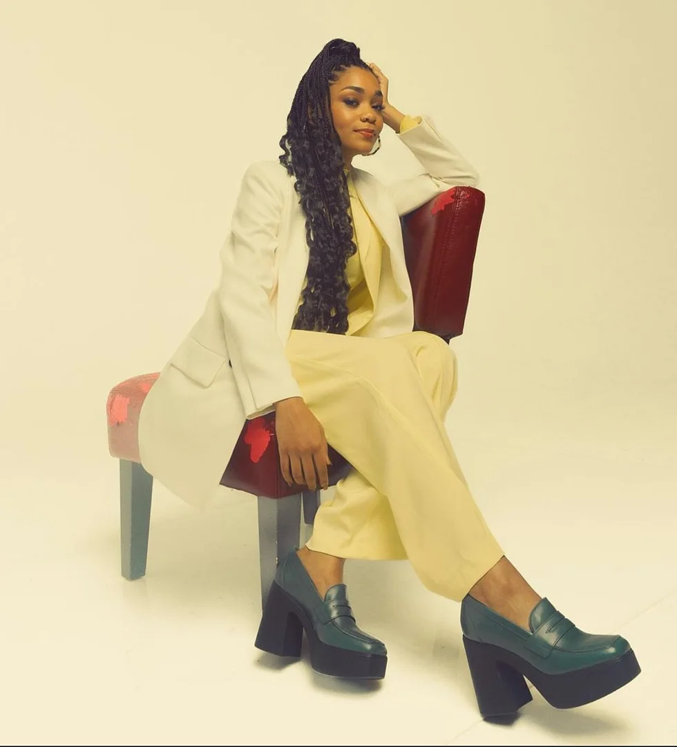 Portrait of an adult woman with braided hair wearing a white blazer and yellow pants, seated on a red chair against a white backdrop.