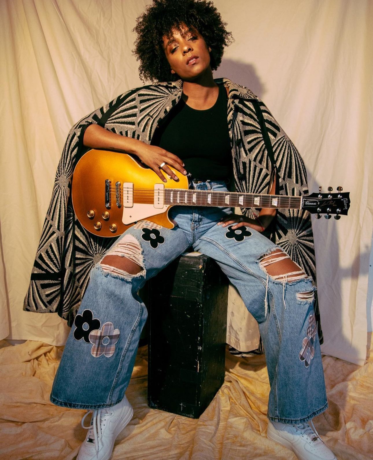 A stylish woman with curly hair poses with a gold electric guitar, wearing patterned denim jeans and jacket against a neutral backdrop.