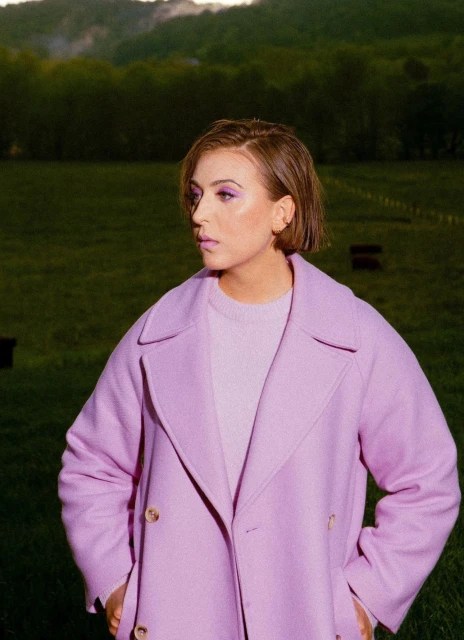 Woman in lilac coat stands outdoors in field, looking left with hills and trees in the background under soft daylight.