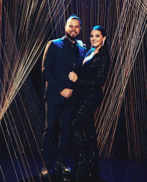A smiling couple poses indoors: the man in a suit with a bolo tie and the woman in a sequined gown, standing against a stringed backdrop.