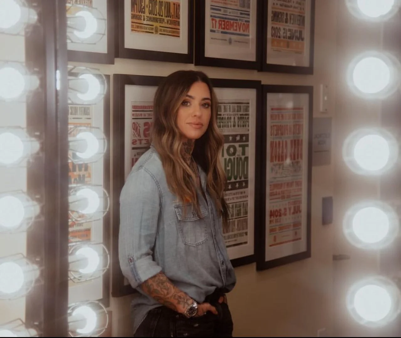 Woman with long wavy hair wearing a denim shirt posing indoors, standing next to a wall with framed posters and light bulbs.