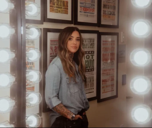 Woman with long wavy hair wearing a denim shirt posing indoors, standing next to a wall with framed posters and light bulbs.