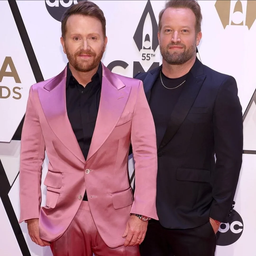 Two adult males pose on a red carpet at an awards show. One wears a pink suit, the other a dark suit. Logos and text are in the background.
