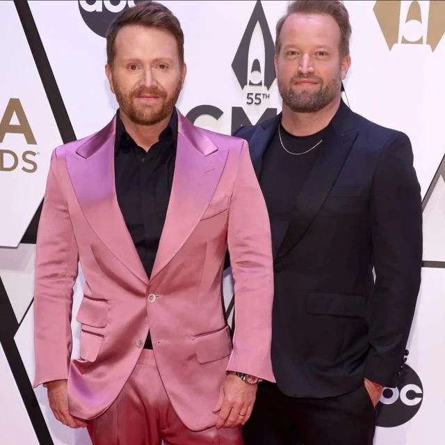 Two adult males pose on a red carpet at an awards show. One wears a pink suit, the other a dark suit. Logos and text are in the background.