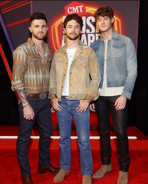 Three young adult males pose on a red carpet for the CMT Music Awards, wearing western-style jackets and boots.