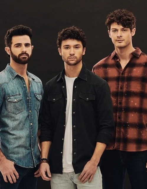 Three adult males stand indoors against a dark backdrop wearing denim and plaid shirts, looking directly at the camera.