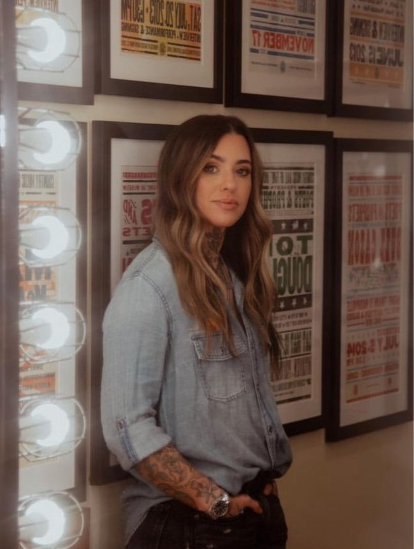 Portrait of a young adult woman with tattoos in a denim shirt, posing in front of framed posters and a wall of vanity lights.