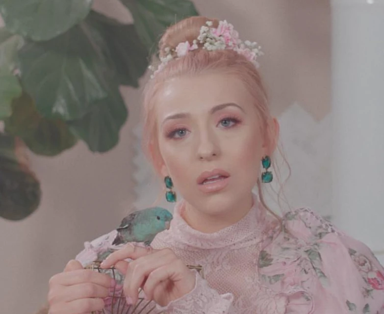 Woman with pink hair, wearing floral dress and flower crown, holding a turquoise bird in an indoor setting with soft lighting.