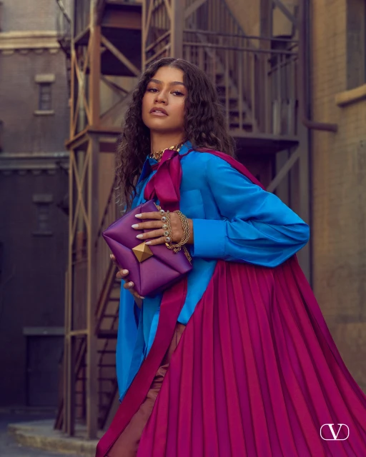 Zendaya poses in a fashion editorial image with a blue blouse, fuchsia skirt, gold jewelry, and a purple purse in an urban setting.