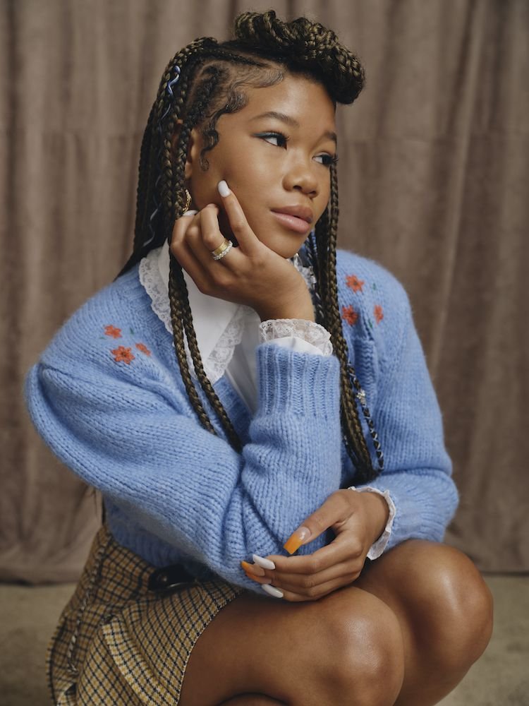 Portrait of a young woman with braided hair, wearing a light blue cardigan with floral details, posing indoors against a neutral backdrop.