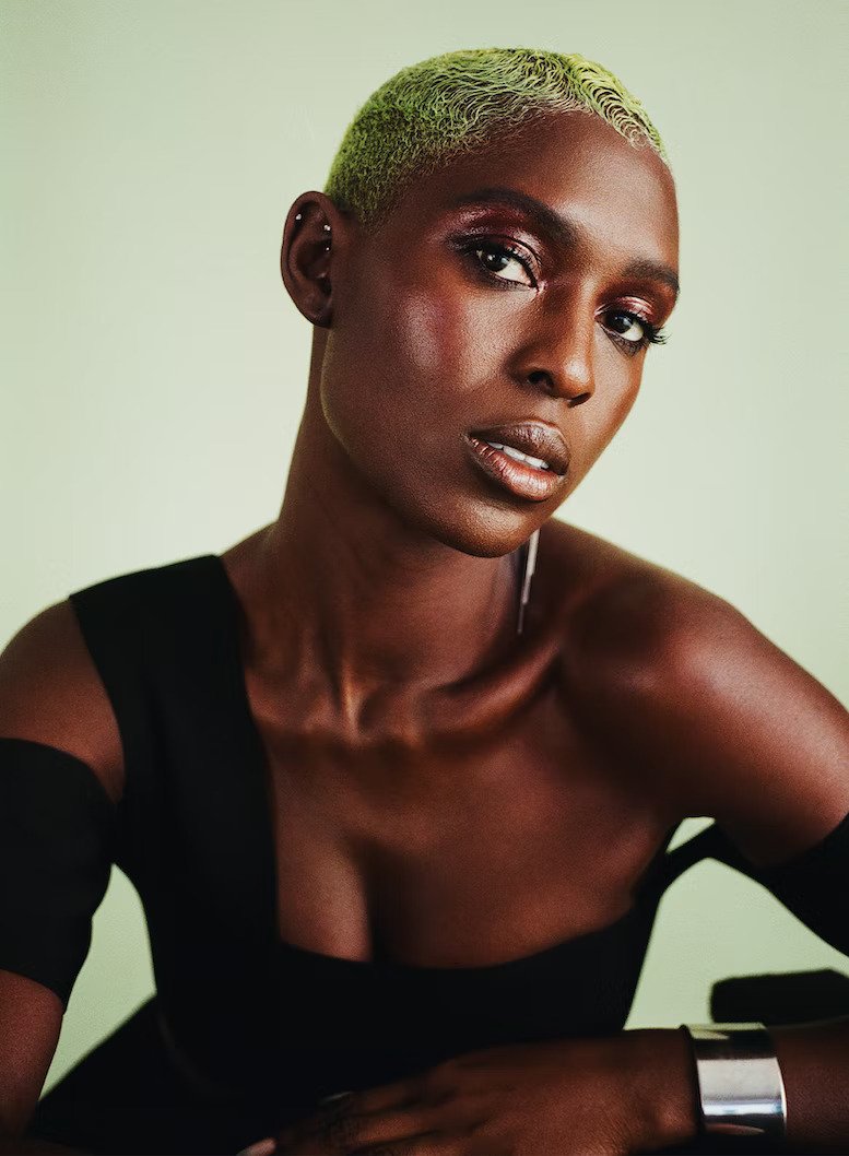 Portrait of a stylish black woman with short green hair in a black dress with a silver cuff, set against a soft green backdrop.