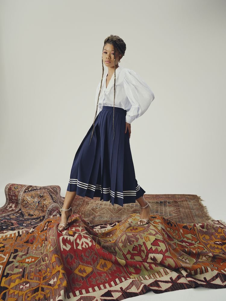 A young Black woman poses in a white and blue blouse and navy skirt, stepping across a pile of oriental rugs in a studio setting.