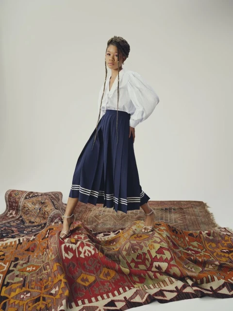 A young Black woman poses in a white and blue blouse and navy skirt, stepping across a pile of oriental rugs in a studio setting.