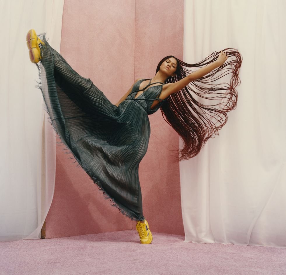 Woman in flowing dress and yellow sneakers strikes dynamic pose against pink and white backdrop in a fashion forward studio shot.