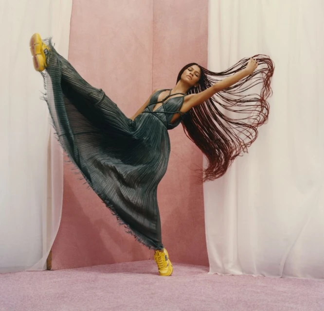 Woman in flowing dress and yellow sneakers strikes dynamic pose against pink and white backdrop in a fashion forward studio shot.