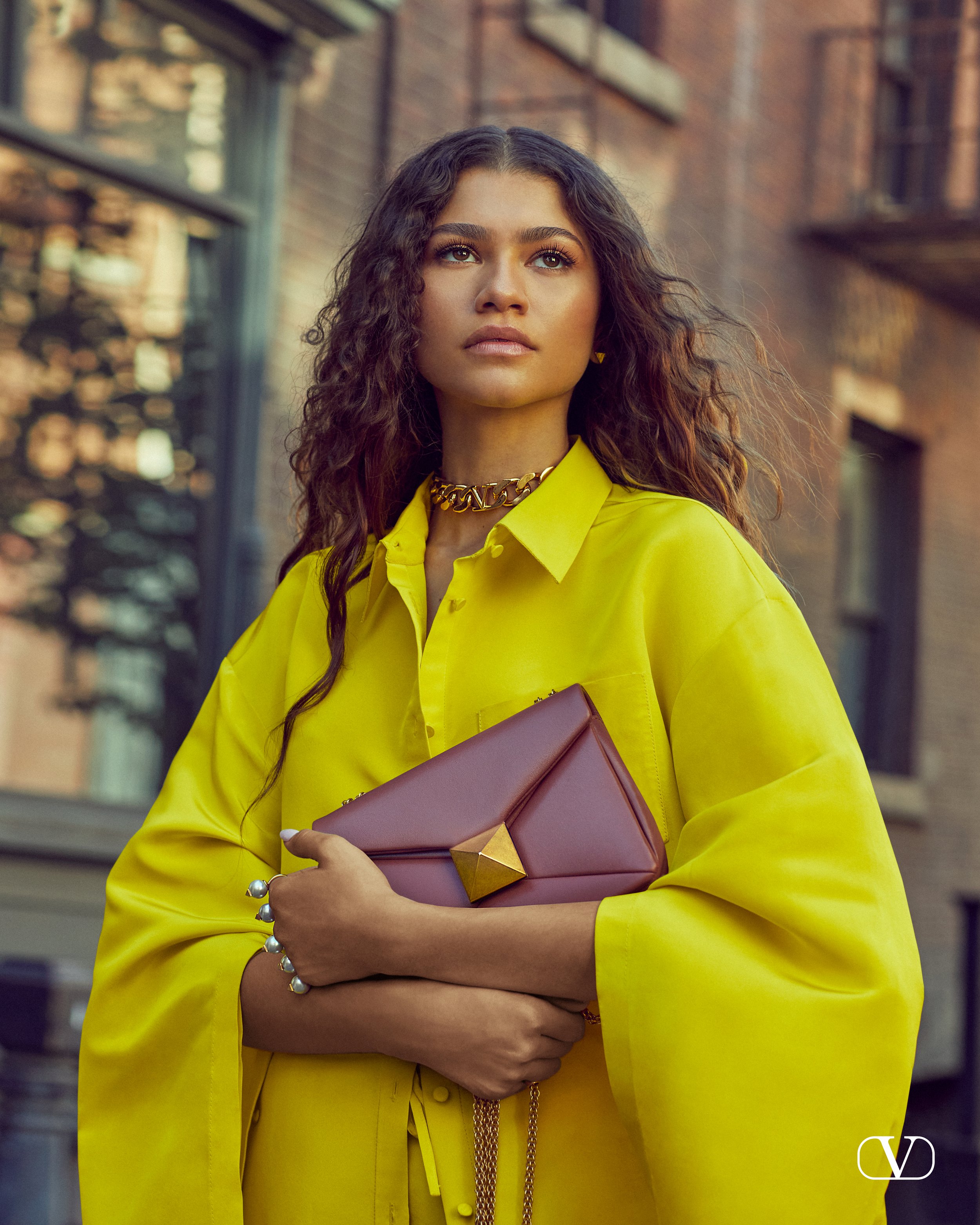 Zendaya is shown in a medium shot, wearing a yellow top and clutching a pink handbag against an urban backdrop of a city street.