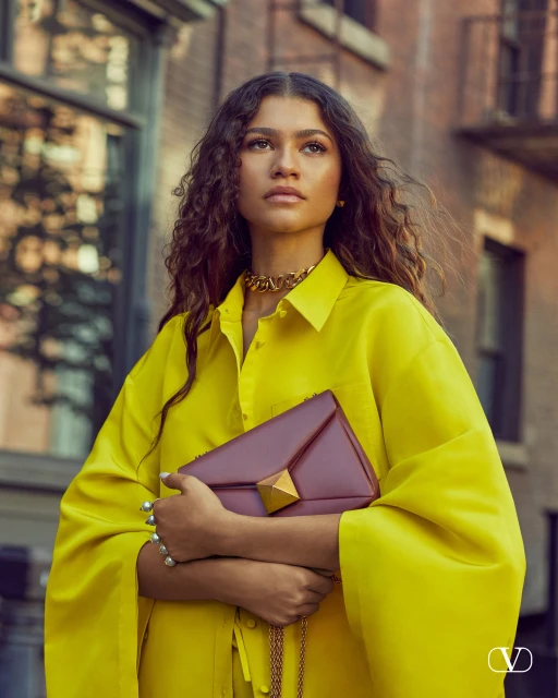 Zendaya is shown in a medium shot, wearing a yellow top and clutching a pink handbag against an urban backdrop of a city street.