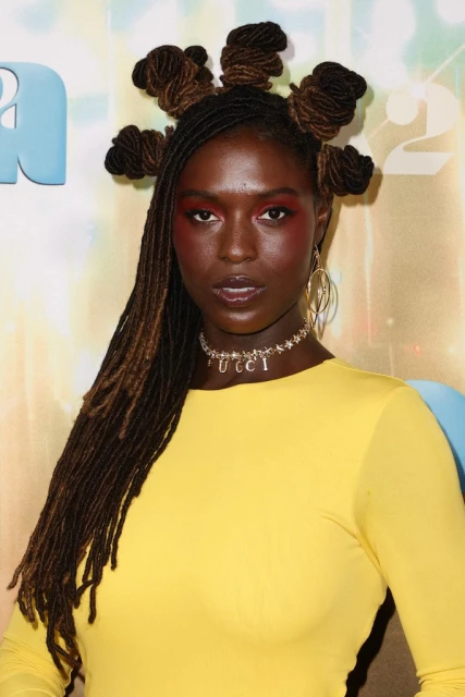 Close-up portrait of an African American woman with creative hairstyle, wearing a yellow top and a necklace, posing against a blurred backdrop.