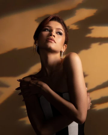 Zendaya posing in a strapless gown with dramatic projected cloud lighting on a warm tan background.