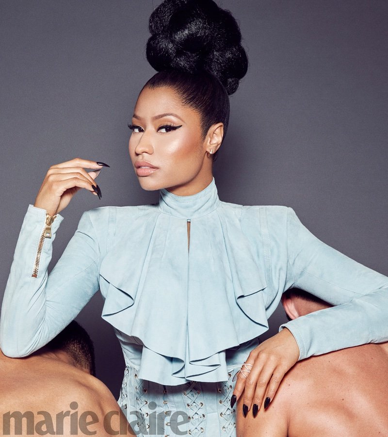 Nicki Minaj poses in a light blue outfit with a high bun hairstyle, leaning on two male figures in a studio setting for Marie Claire.