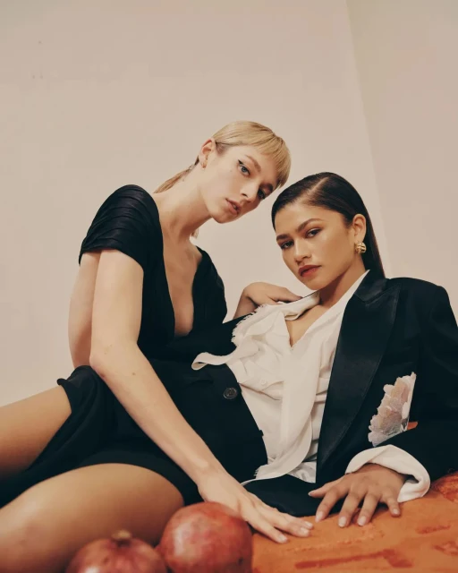 Two women, one with blonde hair in a black dress, the other with dark hair in a blazer, pose together with pomegranates.