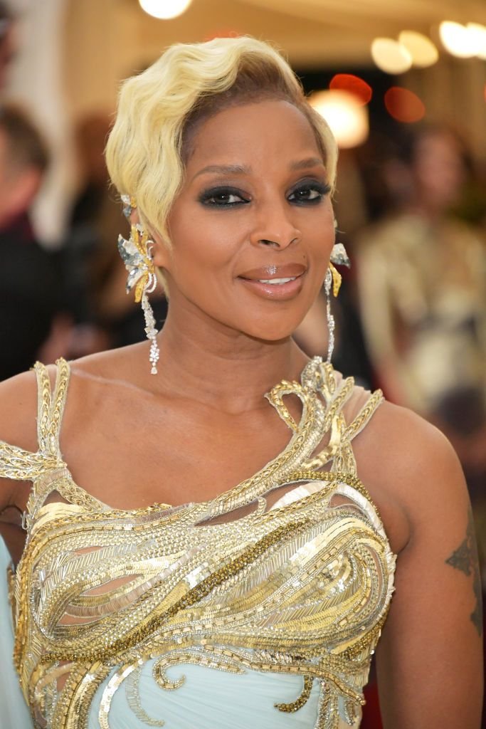 Close-up of Mary J. Blige with blond hair and jeweled gown, smiling softly at the camera. The background is blurred.