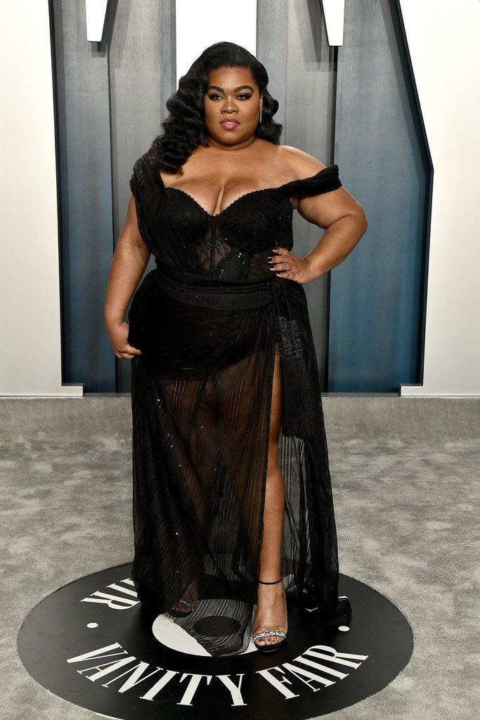 A woman with dark wavy hair wearing an off-the-shoulder black evening gown standing on a Vanity Fair logo round floor disk.