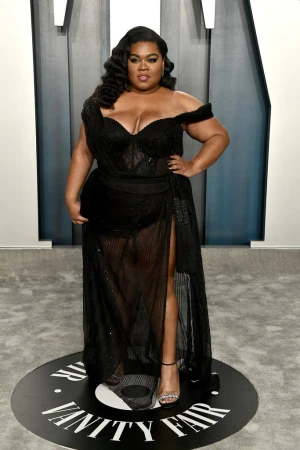 A woman with dark wavy hair wearing an off-the-shoulder black evening gown standing on a Vanity Fair logo round floor disk.